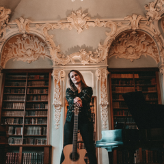 Foto von Sophie Chassee. Sie steht  augestützt auf ihrer Gitarre in einer Bibliothek.in einer Biblio