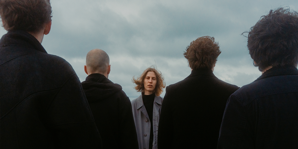 Bandphoto der Whispering Sons. Die Sängerin schaut in die Kamera. Die männlichen Mitglieder haben den Rücken zur Kamera gekehrt und sind ganz in schwarz angezogen.