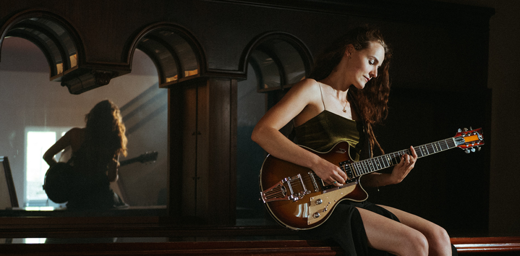 Die Künstlerin sitzend auf einer Bank mit einer E-Gitarre. Im Hintergrund erscheint ihr Spiegelbild