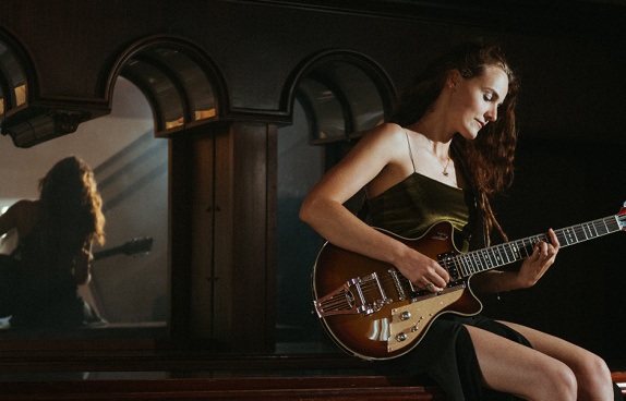 Die Künstlerin sitzend auf einer Bank mit einer E-Gitarre. Im Hintergrund erscheint ihr Spiegelbild
