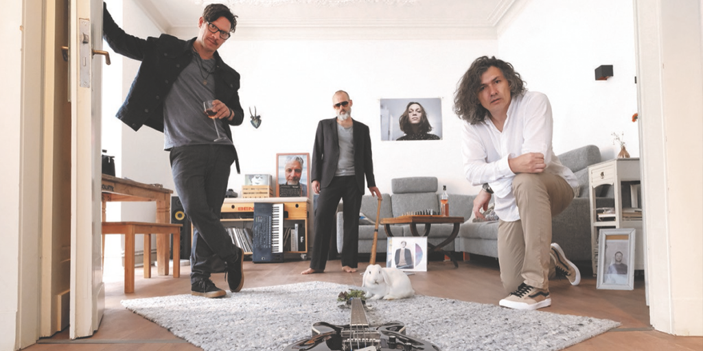 Drei Musiker der Band in einem Wohnzimmer. Drei andere Bandmitglieder sind auf Fotos erkennbar.