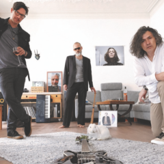 Drei Musiker der Band in einem Wohnzimmer. Drei andere Bandmitglieder sind auf Fotos erkennbar.