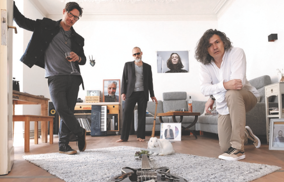 Drei Musiker der Band in einem Wohnzimmer. Drei andere Bandmitglieder sind auf Fotos erkennbar.