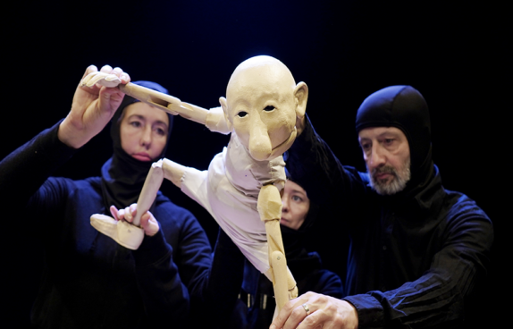 Zwei Marionettenspieler in einem blauen Ganzkörperanzug halten eine weiße Marionette an Armen und Beinen fest.