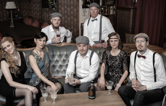 Bandfoto der Shabby Chicks. Fünf Mitglieder sitzen auf einer Ledercouch. Zwei Bandmitglieder stehen hinter dem Sofa
