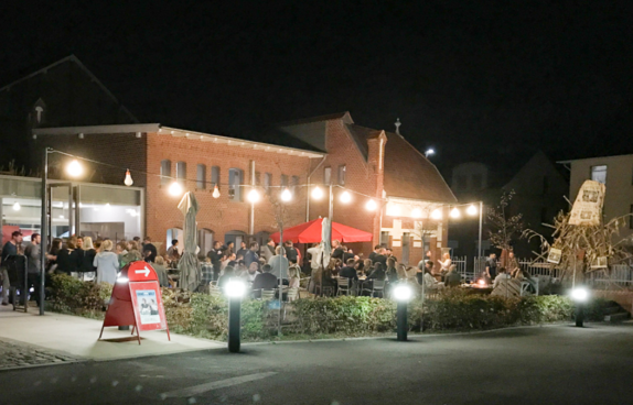 Foto von Bistro außen. Gäste stehen auf der Terrasse. Es ist dunkel.
