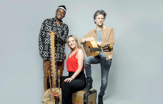 Portrait der Gruppe vor einem grauen Hintergrund. Man sieht die Sängerin Céline Scheen, Mamadou Dramé mit seiner kora und Karim Baggili mit seiner Gitarre.