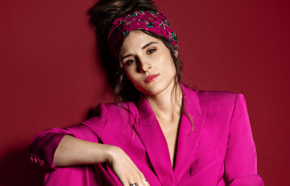 Portrait de l'artiste. Elle porte un blazer rose et un bandeau.
