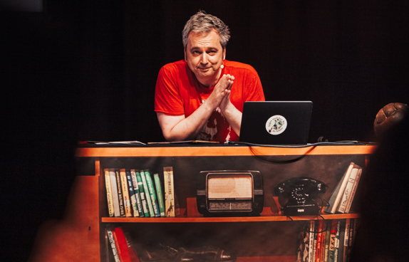Der Künstler Arnd Zeigler sitzt wie in seiner Fernsehsendung an seinem Schreibtisch mit Laptop. Der Schreibtisch hat ein eingebautes Regal und man sieht Bücher und ein altes Radio aus den 70er- Jahren.