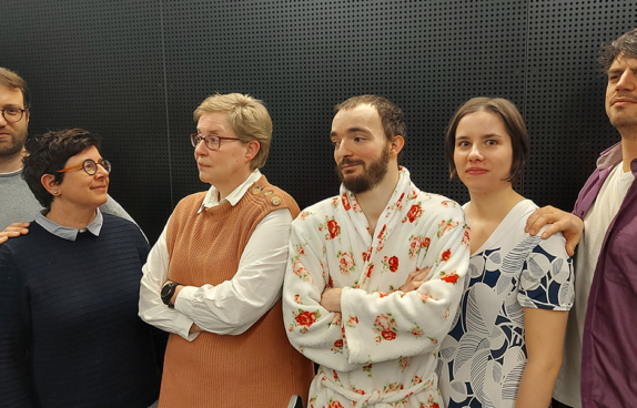 Eine Gruppe von fünf Personen steht vor einer schwarzen, perforierten Wand. Sie schauen in unterschiedliche Richtungen und wirken nachdenklich oder freundlich. Links steht eine Person mit kurzen, dunklen Haaren und Brille, die eine cremefarbene Strickjacke trägt. Daneben eine Person mit kurzem, blondem Haar und einem hellblauen Pullover mit einem Pferdemotiv. In der Mitte steht eine Person mit dunklen Haaren und Bart, die eine rot-weiß-blaue Kapuzenjacke trägt. Rechts daneben ist eine Person mit kurzem, dunklem Haar in einem grauen Hemd. Ganz rechts steht eine Frau mit schulterlangem, braunem Haar und einem grünen Pullover, die lächelt. Die Szene wirkt locker und ungezwungen.