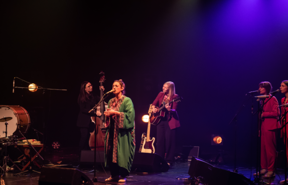Eine Musikgruppe tritt auf einer Bühne unter farbigem Scheinwerferlicht auf. In der Mitte steht eine Sängerin im grünen Gewand am Mikrofon. Links spielt ein Schlagzeuger ein Set aus Trommeln, während eine Musikerin Kontrabass spielt. Rechts neben der Sängerin spielt eine Gitarristin Akustikgitarre. Ganz rechts stehen drei Background-Sängerinnen in roten Anzügen an Mikrofonen. Die Bühne ist in warmes und violettes Licht getaucht, und Instrumente sowie Verstärker sind im Hintergrund sichtbar.