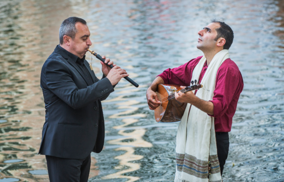 Zwei Musiker spielen traditionelle Instrumente an einem Gewässer. Der Mann links trägt einen schwarzen Anzug und spielt eine Duduk, eine traditionelle armenische Flöte. Der Mann rechts trägt ein weinrotes Hemd mit einer weißen, verzierten Schärpe und spielt eine Saz, ein langhalsiges Zupfinstrument. Er blickt nach oben, während er musiziert. Die reflektierende Wasseroberfläche im Hintergrund verleiht der Szene eine ruhige und stimmungsvolle Atmosphäre.