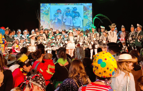 Kinderkarneval auf der Bühne: Eine große Gruppe von Tänzerinnen, Musikern und Karnevalsmitgliedern in grün-weißen Uniformen steht vor einem bunten Publikum. Im Hintergrund läuft eine Projektion mit maskierten Kindern, während viele Gäste in farbenfrohen Kostümen den Auftritt verfolgen.