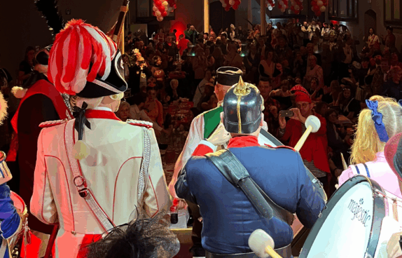 Blick von der Bühne auf ein buntes Karnevalspublikum. Im Vordergrund stehen Musiker und Gardemitglieder in farbenfrohen Uniformen, während das Publikum im festlich geschmückten Saal applaudiert und feiert.
