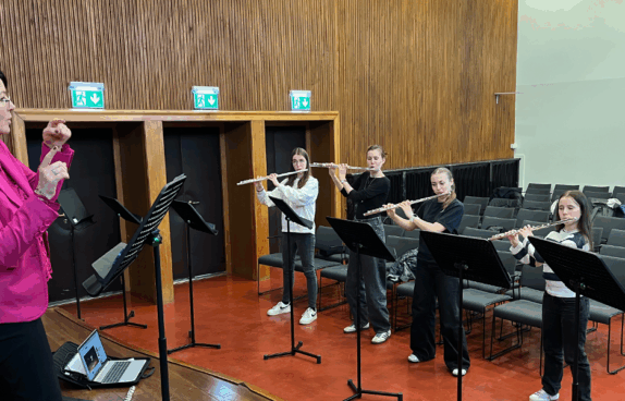 Eine Musiklehrerin dirigiert eine Gruppe junger Flötistinnen während einer Probe in einem Konzertsaal. Die Musikerinnen stehen mit ihren Querflöten vor Notenständern, während im Hintergrund leere Stuhlreihen zu sehen sind.