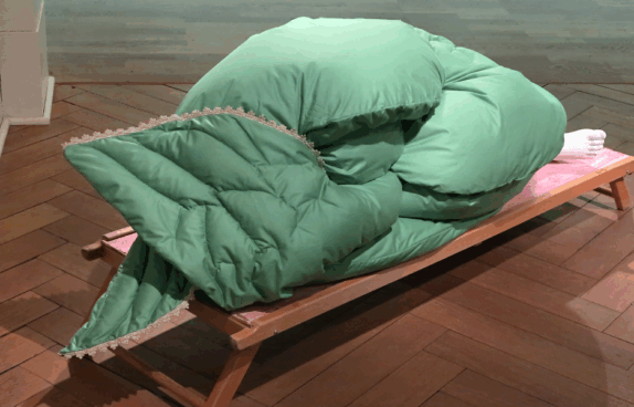 Eine kunstvolle Installation: Auf einer Holzliege liegt eine große, grün gepolsterte Decke, die zu einer kompakten, organischen Form zusammengerollt ist. Am rechten Ende ragen zwei gestrickte, weiße Füße hervor.