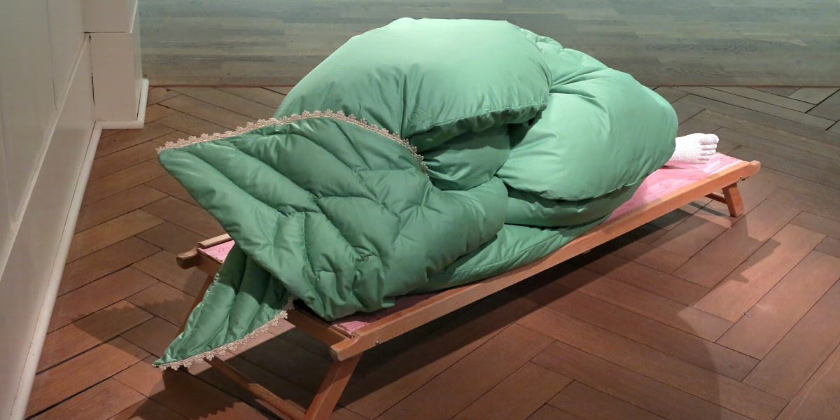 Eine kunstvolle Installation: Auf einer Holzliege liegt eine große, grün gepolsterte Decke, die zu einer kompakten, organischen Form zusammengerollt ist. Am rechten Ende ragen zwei gestrickte, weiße Füße hervor.