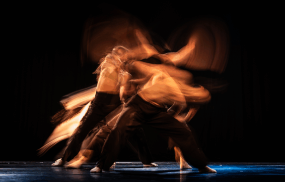 Mehrere Tänzer:innen bewegen sich dynamisch auf einer dunkel beleuchteten Bühne. Durch eine lange Belichtungszeit wirken ihre Körper verschwommen und mehrfach überlagert, sodass fließende, abstrakte Bewegungsbahnen in warmen Orange- und Brauntönen entstehen. Die Szene vermittelt Energie, Rhythmus und Bewegungsintensität vor schwarzem Hintergrund.